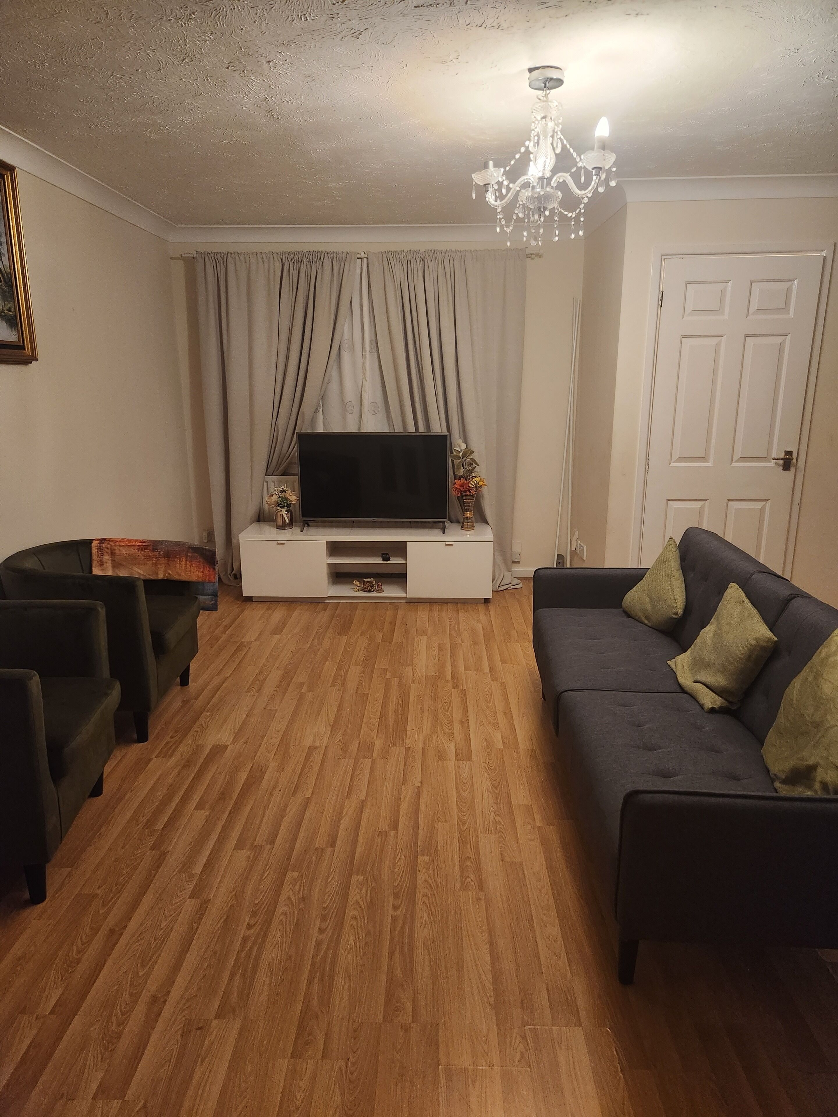 Ein Wohnzimmer mit einem Flachbildfernseher auf einem weißen Schrank, einem dunklen Sofa mit grünen Kissen, einem Holzboden und einer Kristallleuchter.