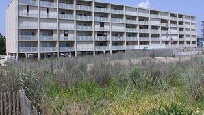A multi-story building with balconies, a parking area in front, and a grassy area in the foreground. - Beachmark 4K-Oceanfront 73rd St, WIFI, Pool, Elev, W/D, AC (Ocean City)