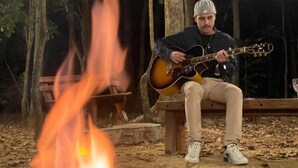 A person sitting on a bench playing a guitar near a campfire in a wooded area. - Recanto das Videiras (Itapira)