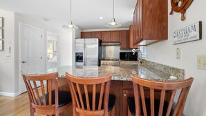 A kitchen with wooden cabinets, a stainless steel refrigerator, a stove, and a granite countertop. There are three wooden chairs with black cushions. - Wonderful dog friendly rental with access path to a private beach deeded at White's Pond! (Chatham)