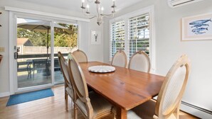 A dining room with a wooden table, six chairs, a chandelier, and a view of the outdoors through large windows. - Wonderful dog friendly rental with access path to a private beach deeded at White's Pond! (Chatham)