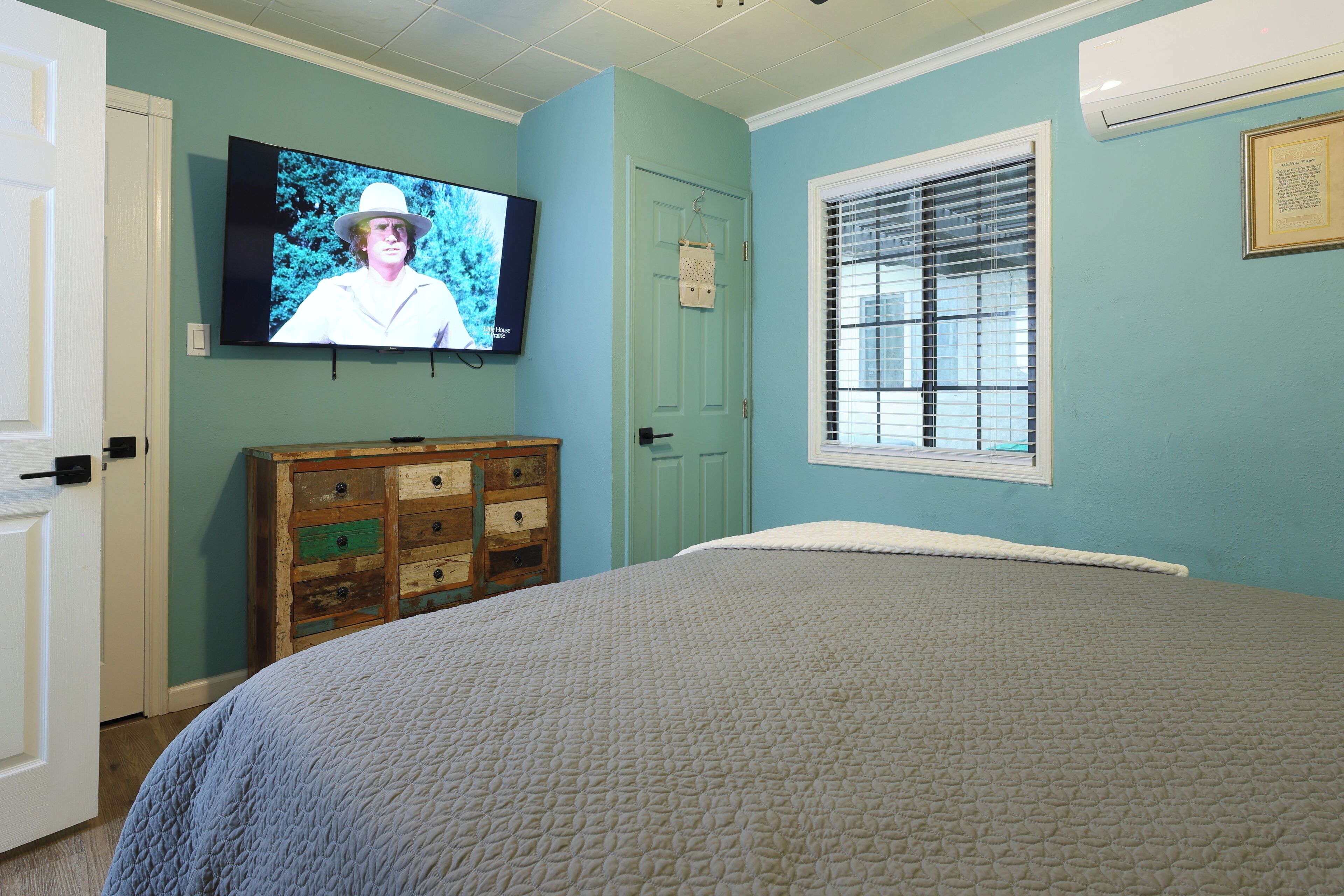 Een slaapkamer met een bed, een dressoir, een aan de muur bevestigde televisie en een raam met jaloezieën.