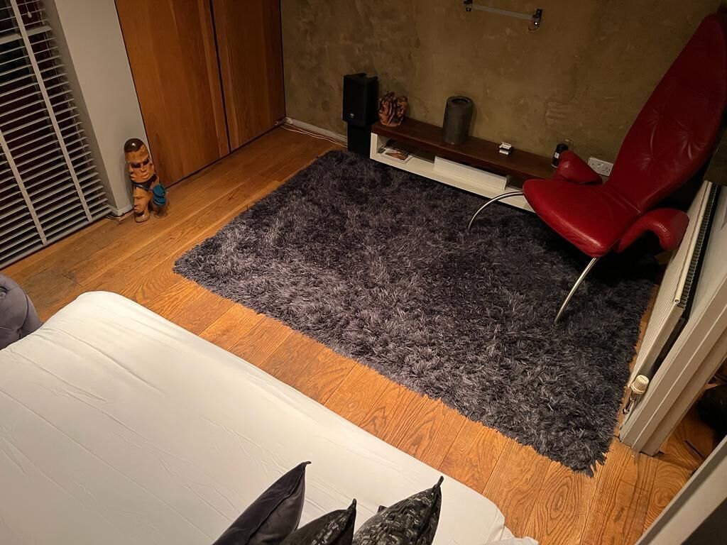 A room with a bed, a red chair, a dark rug, wooden flooring, a wooden wardrobe, and a small toy figure.