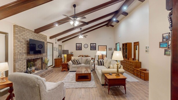 A spacious living room with a fireplace, wooden beams, a ceiling fan, and a television mounted on the wall.