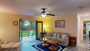 A living room with a sofa, two side tables, a coffee table, a ceiling fan, and a door leading to an outdoor area with chairs.