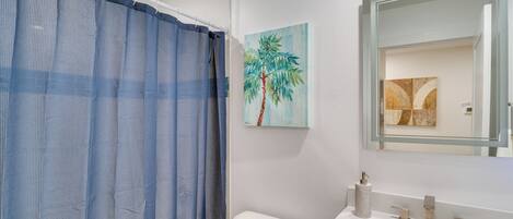 Une salle de bain avec un lavabo blanc, des toilettes, un rideau de douche bleu et un tableau représentant un palmier.