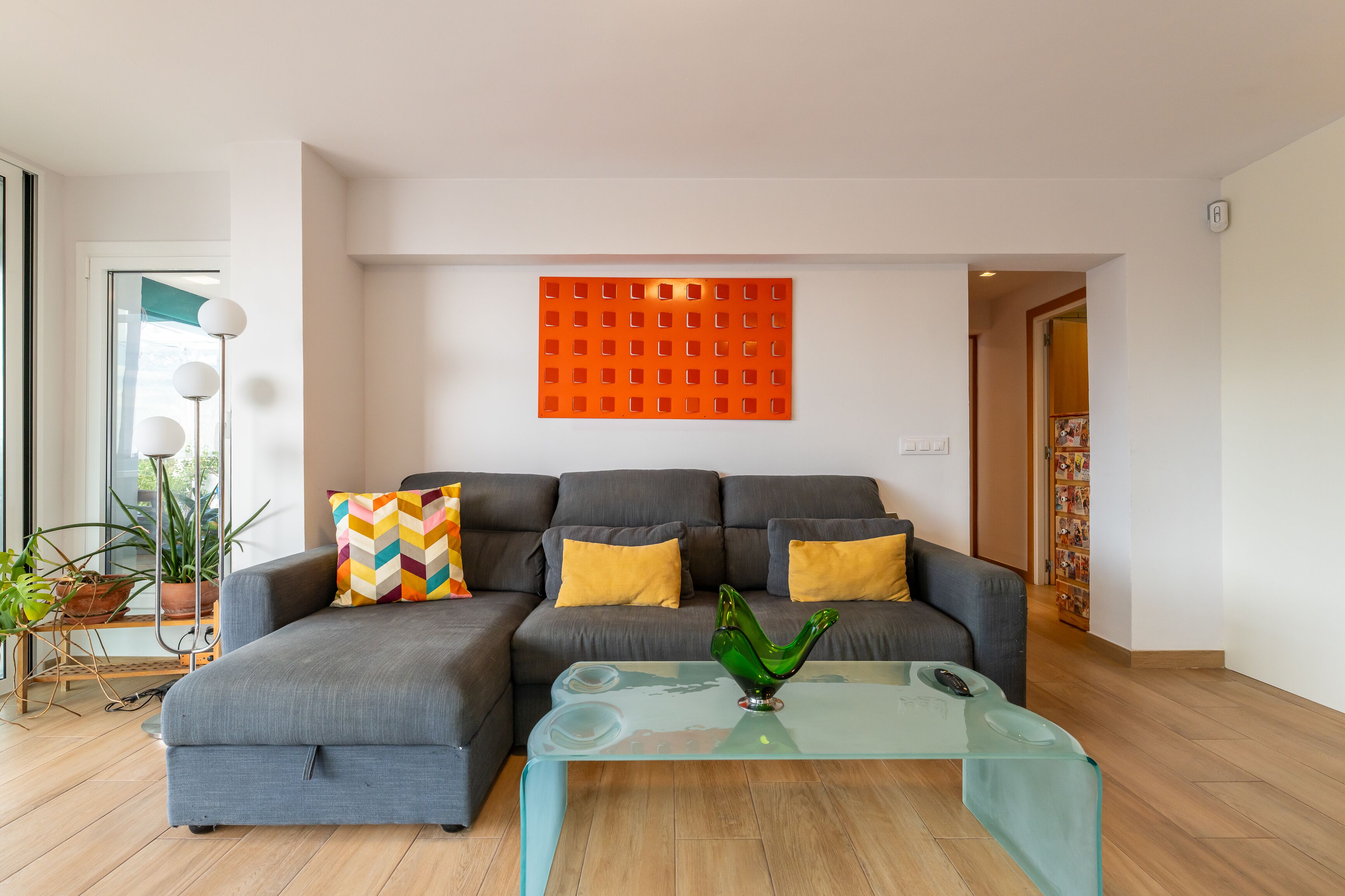A modern living room with a grey sectional sofa, a glass coffee table, and a vibrant orange wall art piece.