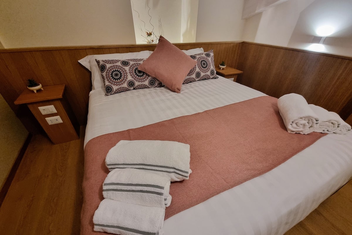 A neatly made bed with a pink blanket, white pillows, and a patterned headboard. There are two bedside tables with a small plant on one.