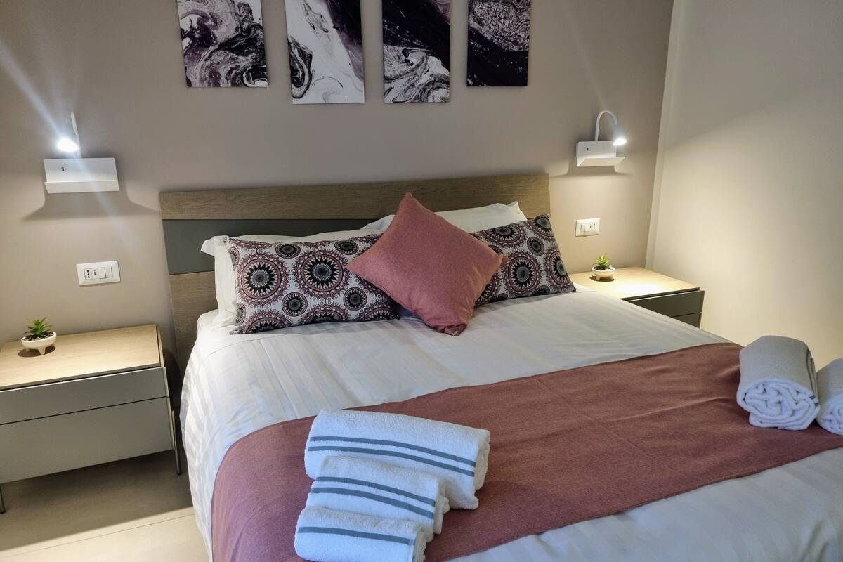 A neatly made bed with patterned pillows, a bedside plant, and a wall-mounted light fixture.