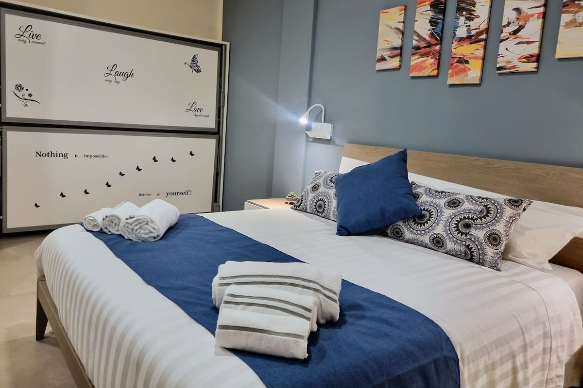 A neatly made bed with blue and white bedding, a blue pillow, and a white pillow with a pattern. There are rolled towels on the bed. The room has a light blue wall and a white ceiling.