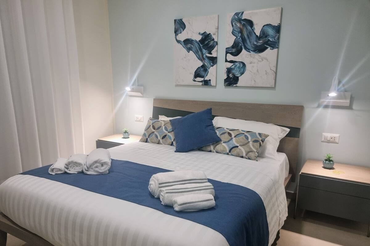 A neatly made bed with blue and white bedding, two bedside tables with lamps, and two wall-mounted paintings.