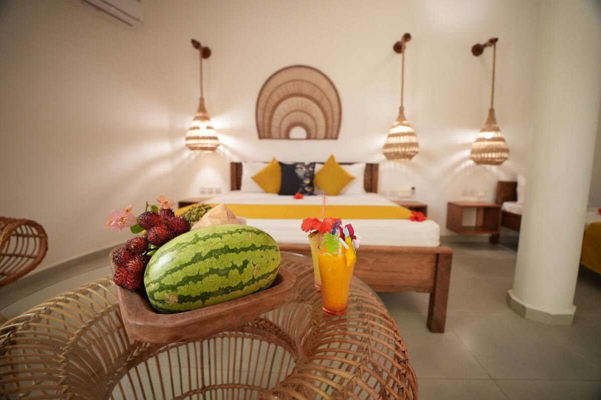 Ein Zimmer mit einem Bett, einem Nachttisch aus Holz mit Obst und einem Getränk, Korbmöbeln und Pendelleuchten.