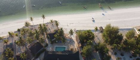 Vista aérea de un resort en la playa con piscina, palmeras y embarcaciones en el agua.