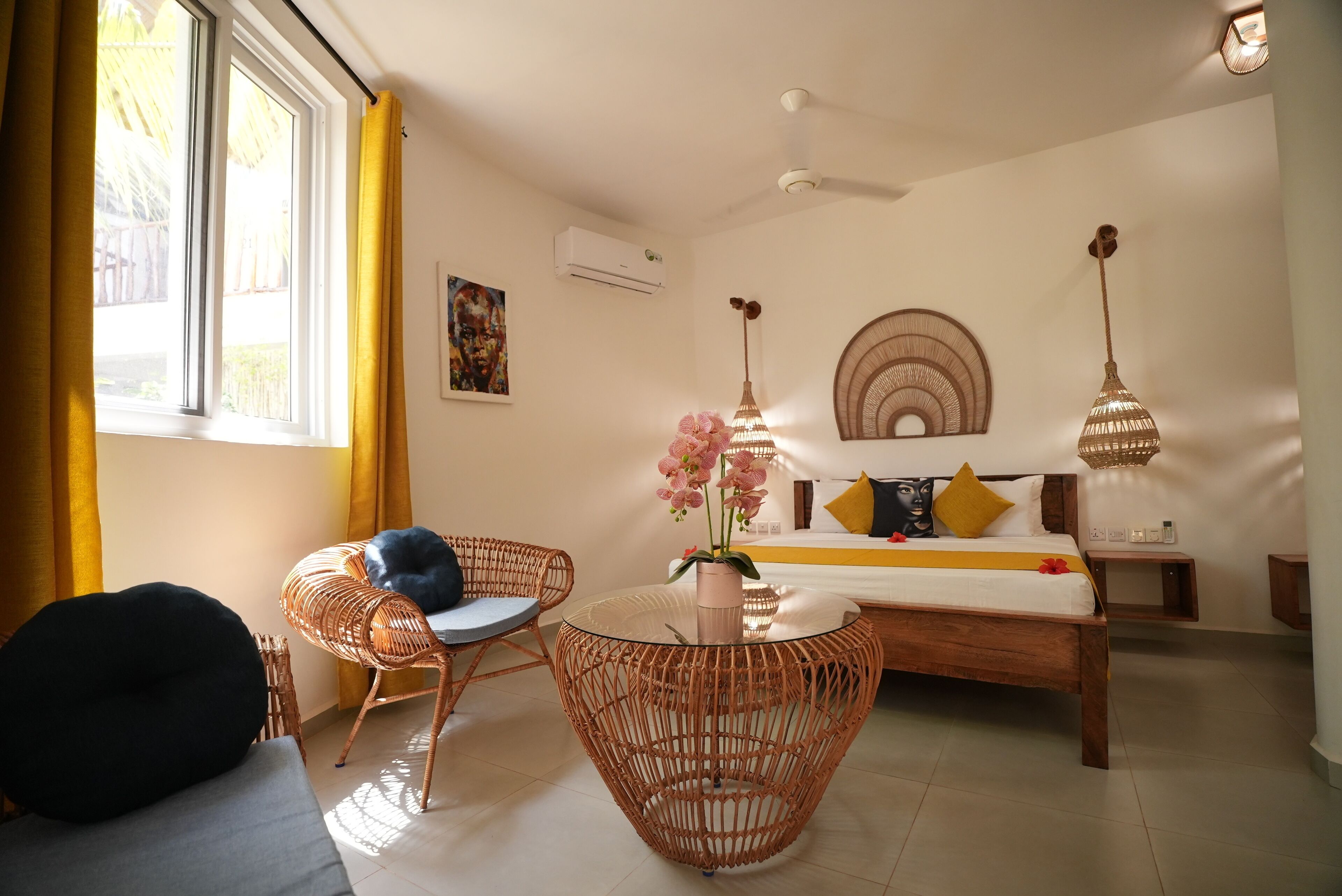 Une pièce comprenant un lit, une table ronde en osier, une chaise en osier, une petite table ronde ornée d’un vase de fleurs et un ventilateur de plafond.