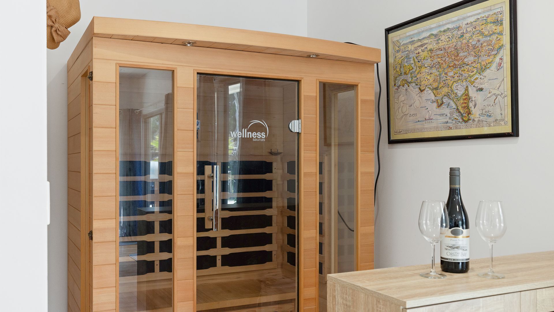 A sauna with a wooden exterior and glass doors, a framed map on the wall, and a bottle of wine on a table.