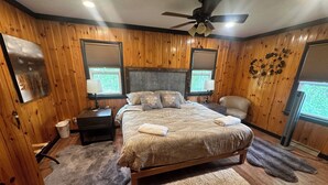 Een slaapkamer met een houten wandpaneel, een bed met een beige dekbedovertrek, twee nachtkastjes met lampen, een plafondventilator en een raam met jaloezieën.