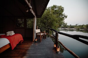Une terrasse en bois avec un lit et un coin salon, donnant sur une étendue d’eau calme, le tout entouré d’arbres.