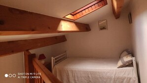 Una habitación pequeña y acogedora, con techo inclinado y vigas de madera, una cama individual, una mesita de noche, una lámpara y un ventanal.