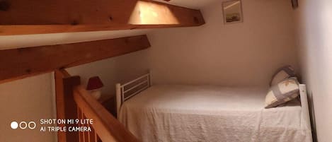 A small, cozy room with a slanted ceiling and wooden beams, a single bed, a nightstand, a lamp, and a small window.