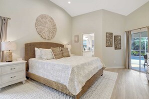 A bedroom with a large bed, a nightstand, a wall-mounted decorative piece, and a view through a glass door. - Live Like a Local:  Charming Pool Home in Berkshire Lakes (Naples)