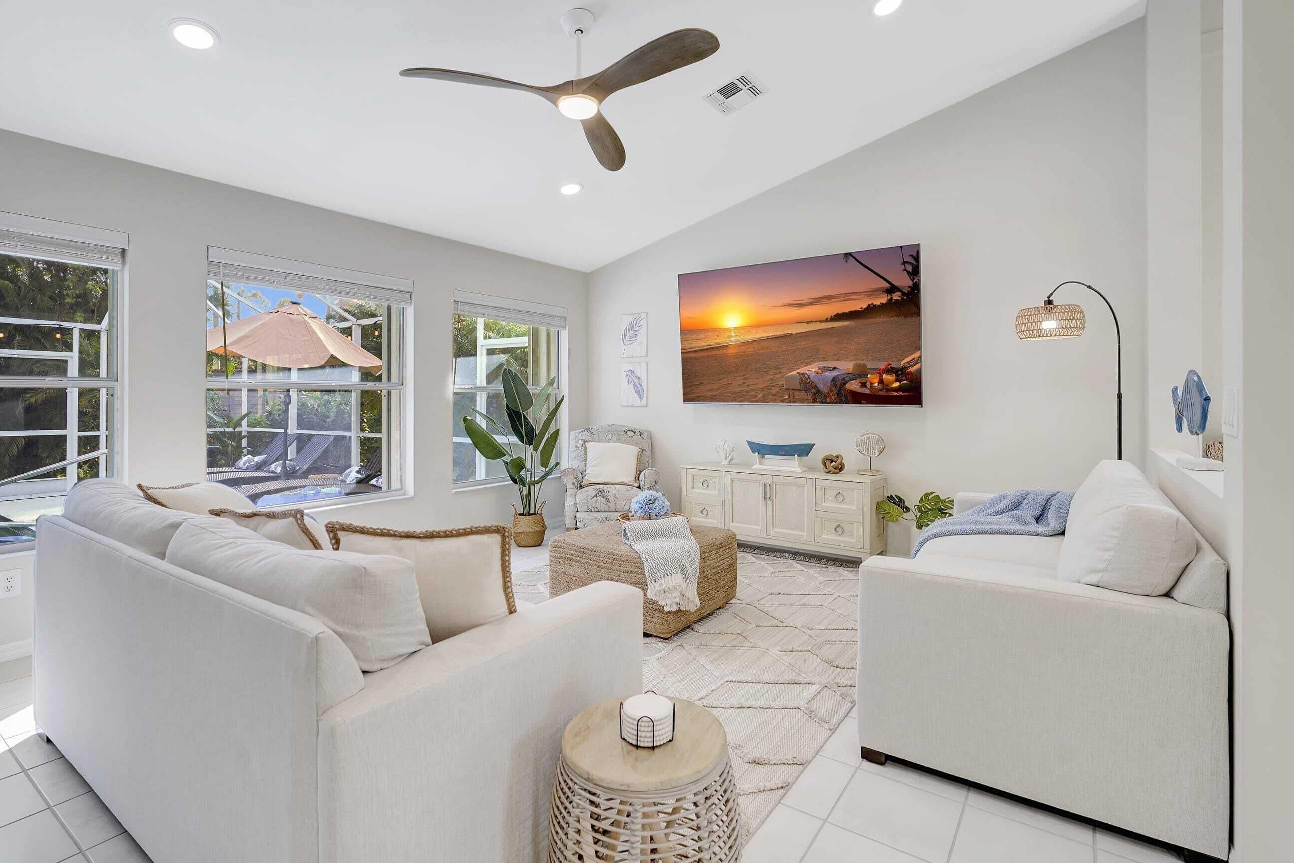 A spacious living room with a large window, a ceiling fan, and a wall-mounted TV displaying a sunset scene.