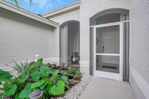 A building with a white exterior, a covered entryway, and a garden with large green plants and rocks. - Live Like a Local:  Charming Pool Home in Berkshire Lakes (Naples)