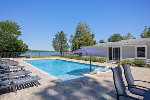 The property features a spacious pool deck with loungers and a lakefront setting, surrounded by mature trees and bordered by scenic water views.