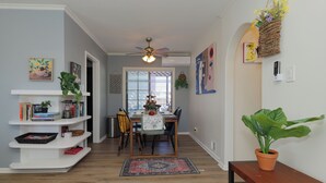 Een eetgedeelte met een houten tafel, stoelen en een plafondventilator. Er is een boekenkast met boeken en decoratieve objecten, en een bloempot op een bijzettafeltje.