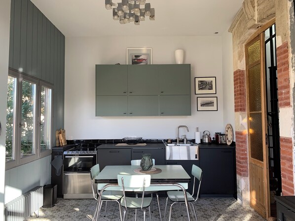 A modern kitchen with a built-in stove, a dining table with chairs, and a large window. - Cosy Little House &garden (Arcachon)
