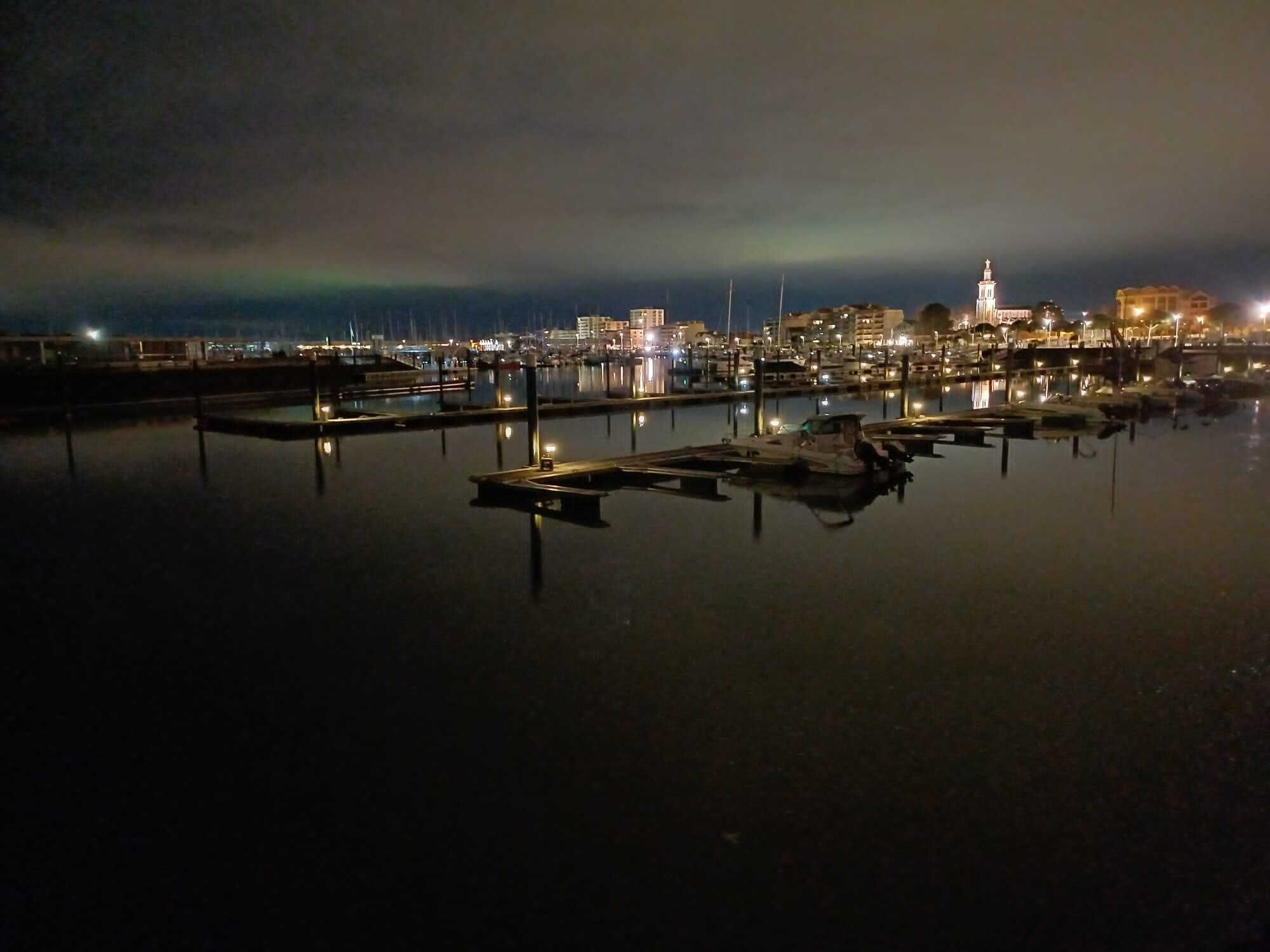 Una marina por la noche con barcos amarrados, agua tranquila y el horizonte de una ciudad al fondo.