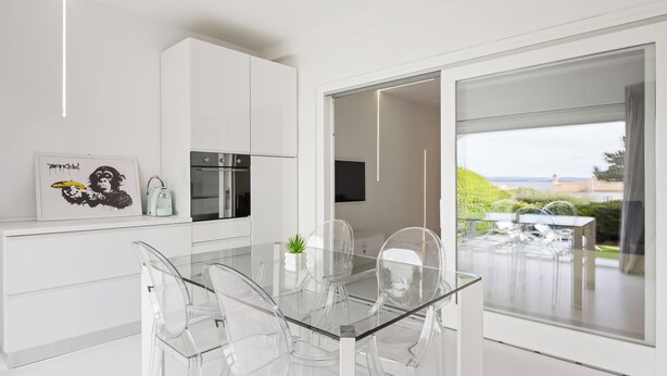 A modern kitchen with white cabinetry, a glass dining table with clear chairs, and a view of the outdoors through a large sliding glass door.