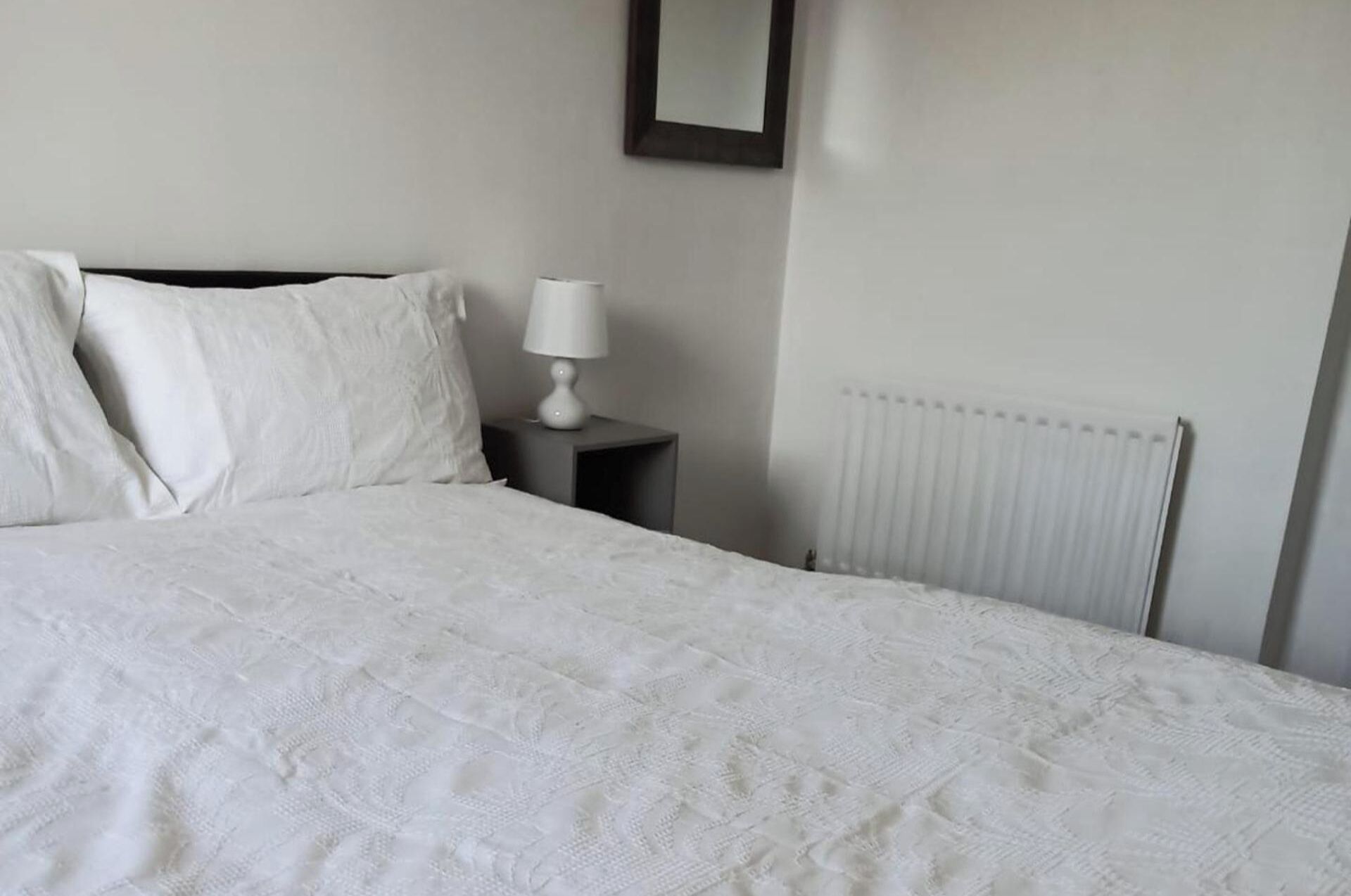 Un lit bien fait, avec du linge de lit blanc, dans une chambre sobre agrémentée d’un radiateur blanc et d’une petite table de chevet avec une lampe.