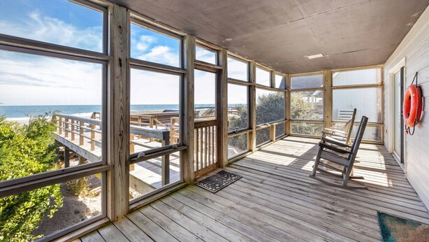 A sunlit room with large windows overlooking a beach, wooden flooring, and a rocking chair. - Beach Nuts - Oceanfront, 4-bedrooms (Pawleys Island)