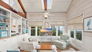 Une pièce avec un plafond en bois, des murs blancs, un ventilateur de plafond, une grande fenêtre donnant sur un beau paysage, un bureau avec un ordinateur et une bibliothèque.