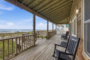 Terraza de madera con vista a la playa, mobiliario exterior y una zona cubierta con barandilla.
