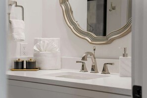 Un baño moderno con un lavamanos blanco, dos grifos, un espejo con marco ondulado y un perchero para toallas.