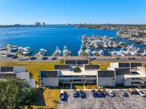 A marina with multiple boats docked, a parking lot with several cars, and a modern building with a flat roof. - Gorgeous Bayfront 2BR 3BA Condo l Marina Views (Panama City Beach)