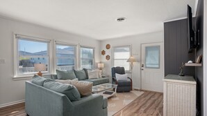 A living room with a grey sofa, a small table, and a chair. There are windows with blinds, a lamp, and a television on a stand. - Direct Beach Access at my newly updated cottage in Nags Head, NC (Nags Head)