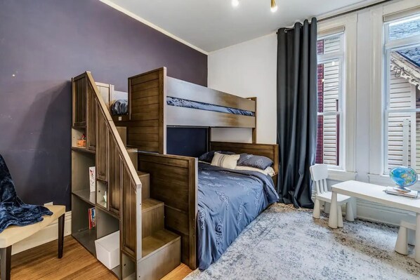 Une chambre à coucher de taille réduite, comprenant un lit superposé, un bureau et une chaise, avec une fenêtre munie de volets et un globe terrestre posé sur le bureau.