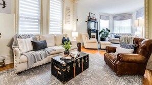 A spacious living room with a beige sofa, a brown leather armchair, and a coffee table with a vintage trunk. - The Victorian on Victoria (Windsor)