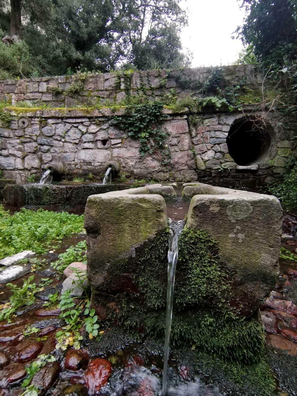 A stone wall with a circular opening, a small waterfall, and moss-covered rocks.