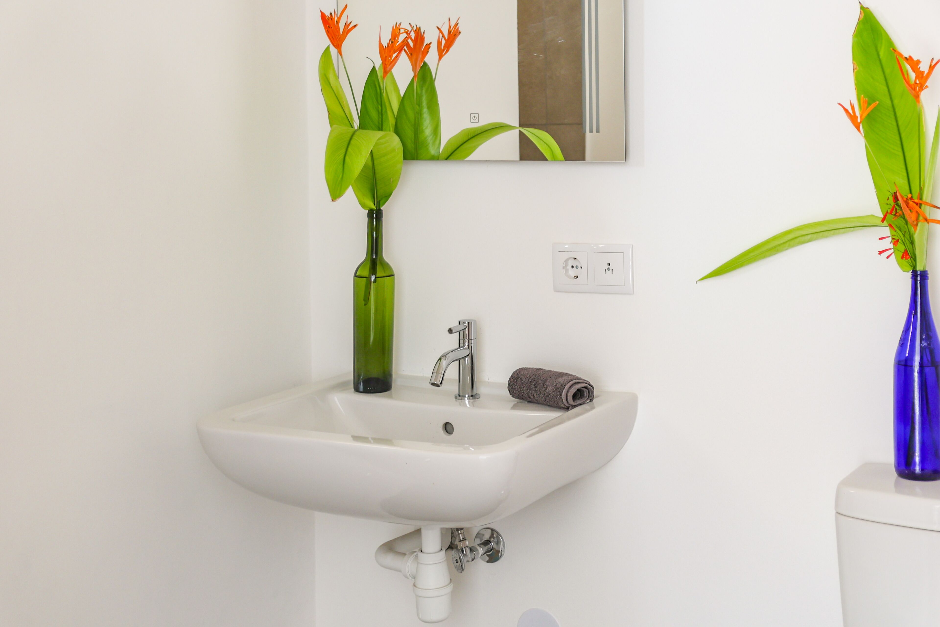 A white bathroom sink with a mirror above it, a green vase with flowers, and a blue vase with flowers.