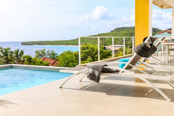 A poolside area with a view of the sea and hills, lounge chairs, and a tiled floor.