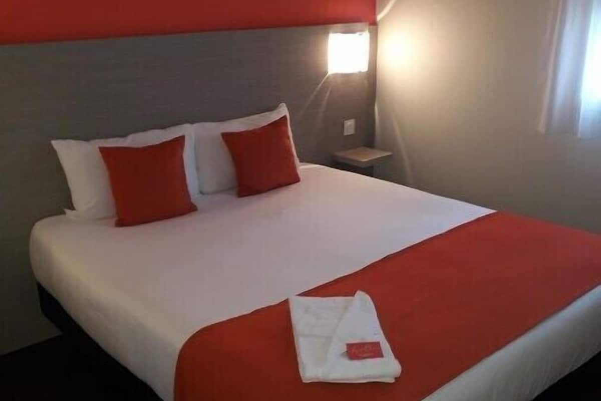 Un lit bien fait, avec une literie blanche et rouge, un plaid rouge et une serviette blanche pliée posée sur le lit.