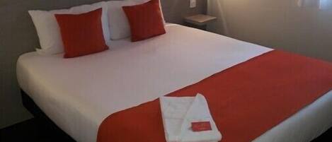 Un lit bien fait, avec une literie blanche et rouge, un plaid rouge et une serviette blanche pliée posée sur le lit.