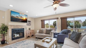 Una sala de estar con chimenea, ventilador de techo y un ventanal con vista a palmeras.