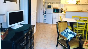 A compact kitchen with white cabinets, a microwave, and a refrigerator. There is a dining area with a table and chairs, and a television on a dresser.