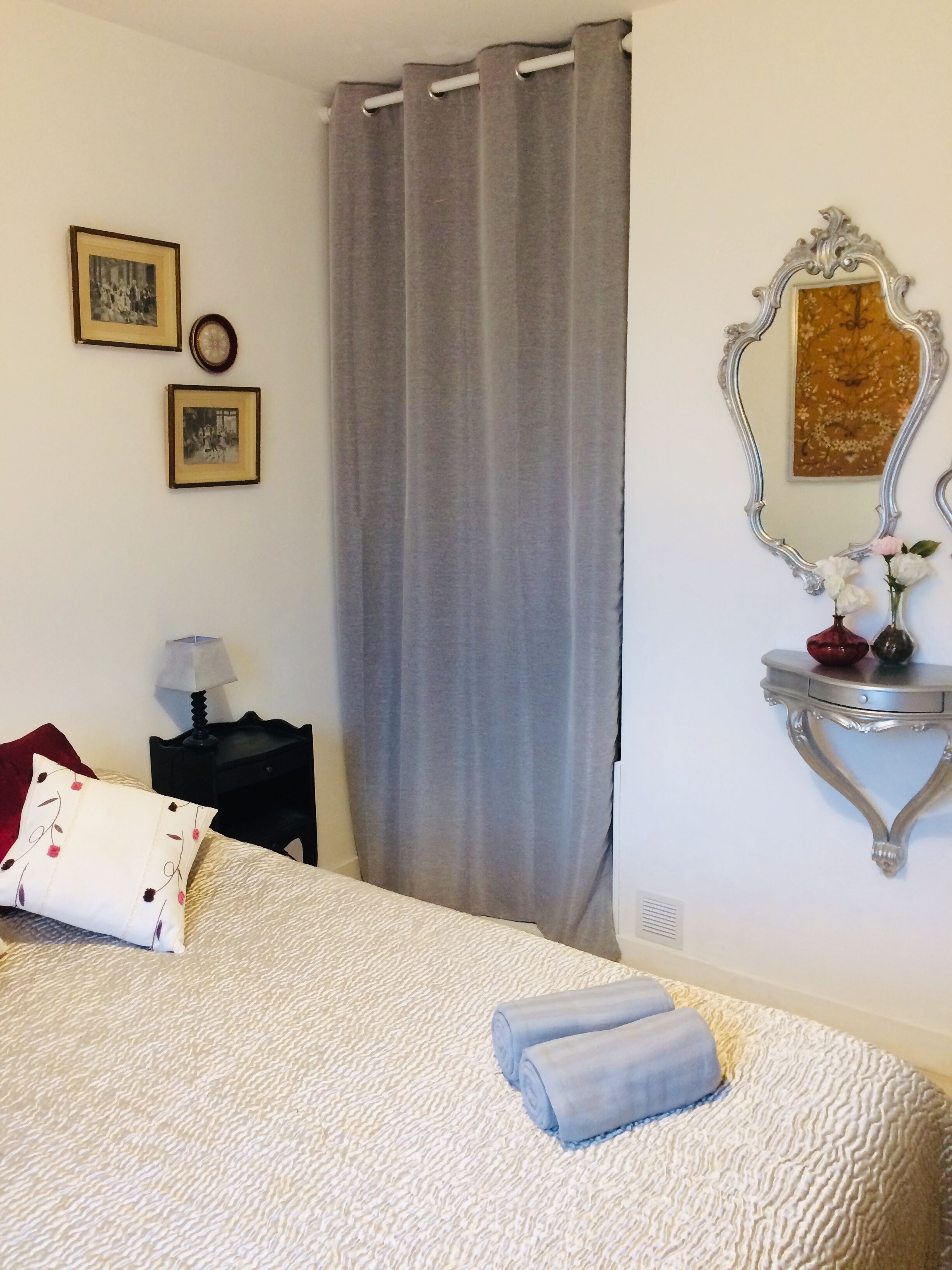 Een net opgemaakt bed met een wit-beige geboeide sprei, een rood kussen en twee opgerolde handdoeken. Een klein nachtkastje met een lamp en een ingelijste foto hangt links. Een groot grijs gordijn bedekt een raam, en een decoratieve spiegel met een bloemenlijst hangt rechts aan de muur.