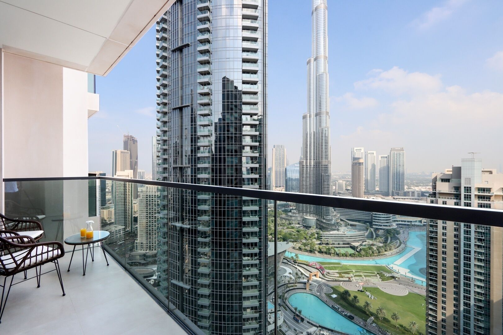 A balcony with a glass railing overlooking a cityscape, including a tall skyscraper and a water feature.