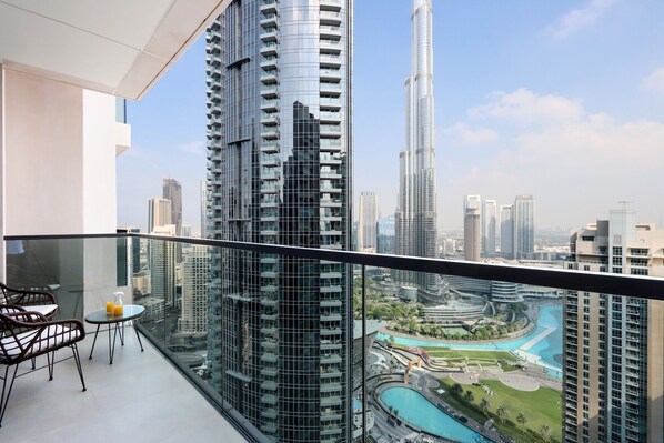 A balcony with a glass railing overlooking a cityscape, including a tall skyscraper and a water feature.